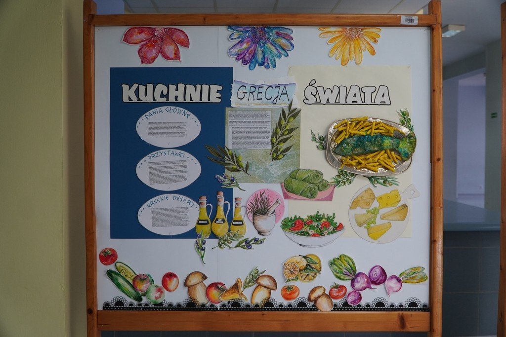 Na zdjęciu widać szkolną tablicę informacyjną z napisem „Kuchnie Świata”. Tablica udekorowana jest kolorowymi, ręcznie wykonanymi ilustracjami potraw i roślin – oliwek, butelek oliwy, sera, sałatki, cytryn, grzybów, warzyw oraz ryby z frytkami. W górnej części znajdują się ozdobne kwiaty, a po lewej stronie umieszczono tekstowe opisy dotyczące dań głównych, przystawek i greckich deserów.