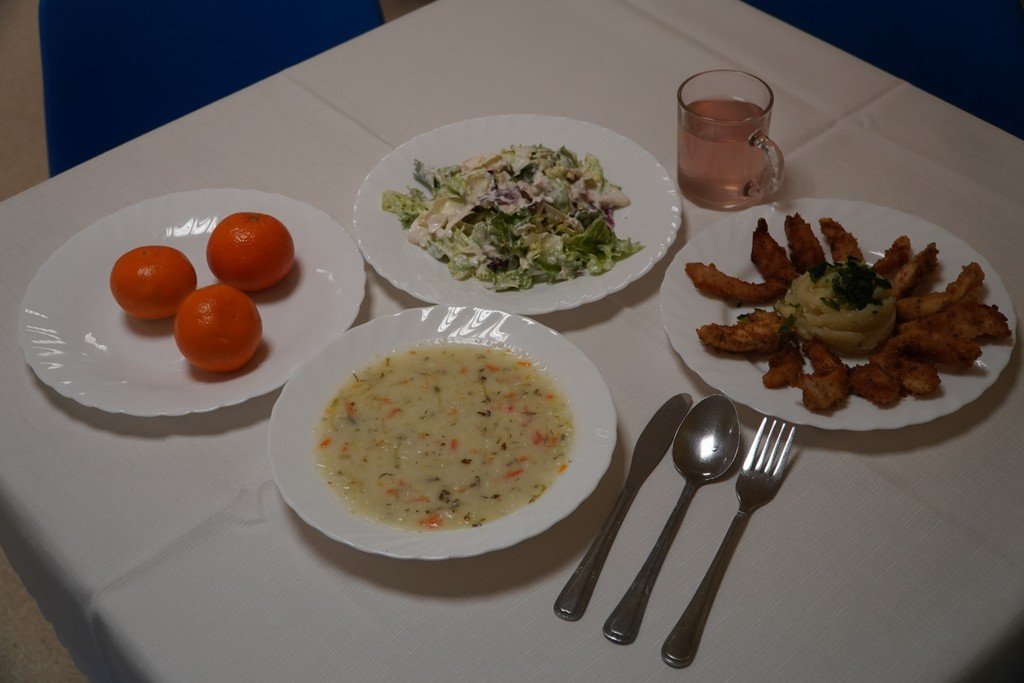 Na zdjęciu widać talerz z panierowaną piersią kurczaka podaną z ziemniakami. Obok znajduje się miseczka zupy ogórkowej z ryżem posypanej świeżą natką pietruszki. Całość uzupełnia porcja chrupiącej sałaty lodowej ze śmietaną i rzodkiewką oraz szklanka kompotu owocowego.