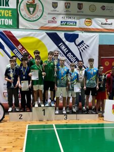 Na zdjęciu widać trzech uczniów Naszej szkoły. Chłopcy, którzy wraz z nauczycielem wychowania fizycznego stoją na podium obok innych zawodników biorących udział w zawodach badmintona w mistrzostwach Polski. Uczniowie mają na szyjach medale, w rękach trzymają puchary i dyplomy. Fotografia została wykonana na hali sportowej podczas uroczystego podsumowania turnieju.