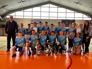 Na zdjęciu widać grupę uczniów oraz nauczycieli, którzy wzięli udział w zawodach sportowych unihokej. Drużyna pozuje do wspólnej fotografii w hali sportowej. Zawodnicy ubrani są w jednakowe, niebieskie koszulki sportowe oraz krótkie spodenki. Na szyjach mają zawieszone medale, jeden z uczniów trzyma w ręku puchar. Część zawodników trzyma również kije do unihokeja, co podkreśla sportowy charakter wydarzenia.