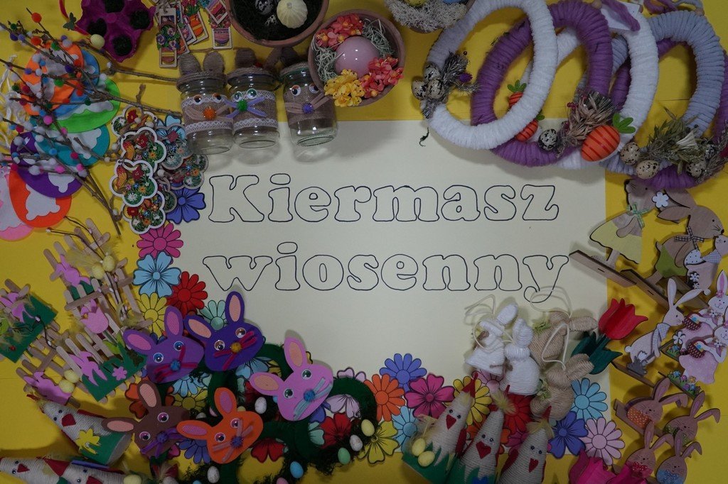 Na zdjęciu znajduje się tablica z dużym napisem „KIERMASZ WIOSENNY”. Dookoła umieszczono różnorodne, ręcznie wykonane, dekoracje o tematyce wiosennej i wielkanocnej, takie jak kolorowe wianki, różnorodne zajączki, zawieszki... Całość utrzymana jest w jasnych, żywych kolorach i sprawia wrażenie bogatej, kreatywnej kompozycji.